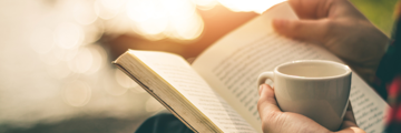 A person sitting outside holding a mug in one hand and a book on their knee.