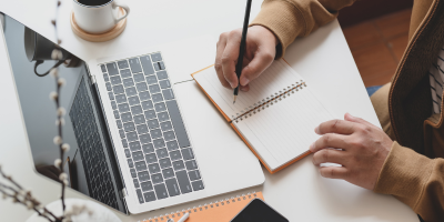 A person at the desk writing notes with the laptop opened