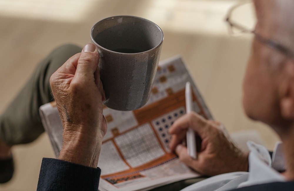 A senior man holding a mug, newspaper and a pen on his lap