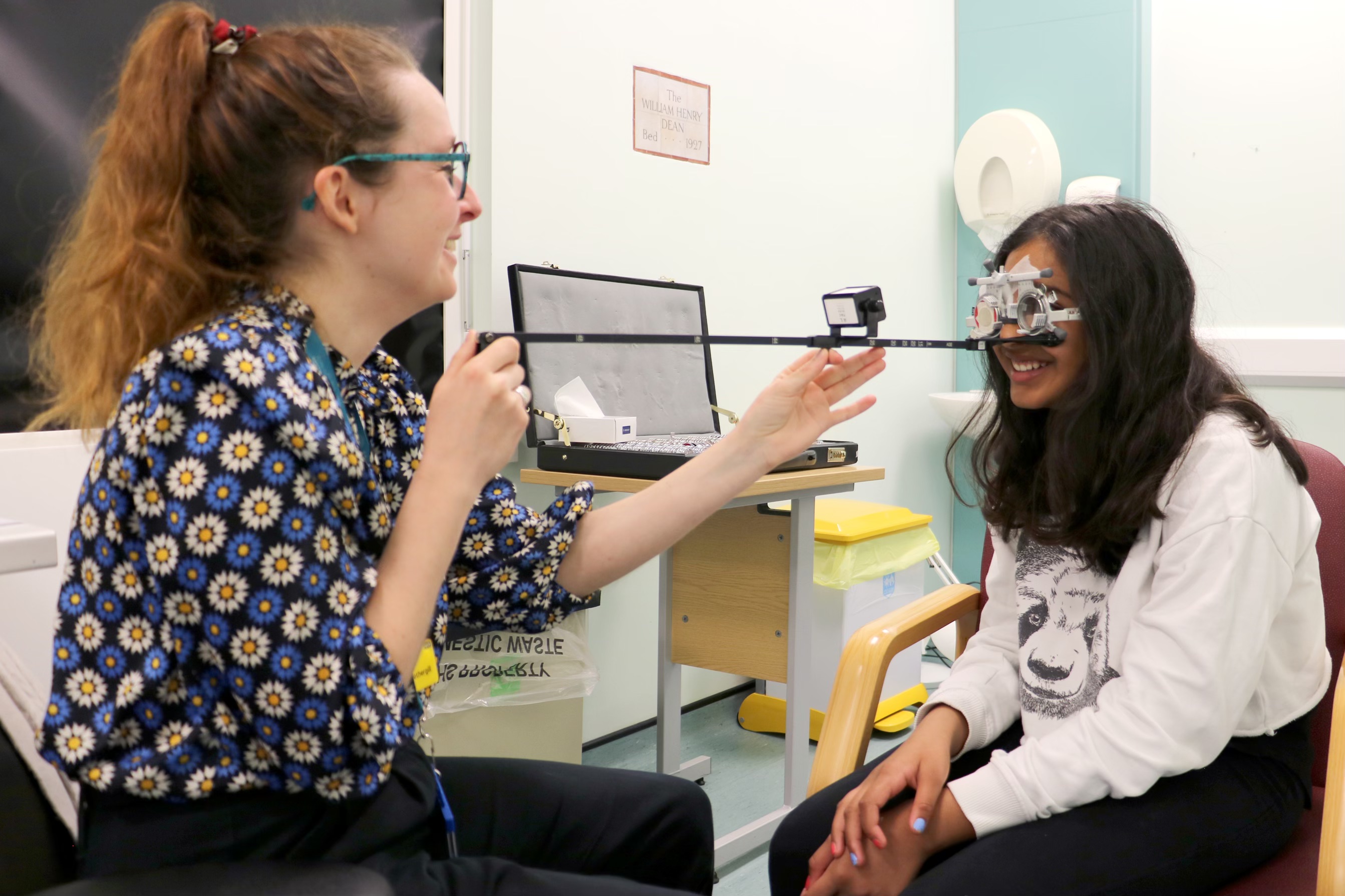 Two people in a consultant room with a child reading letters on a ruler
